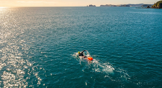 Aerial view of a wetsuit-clad open water swimmer with an orange tow float cutting through sparkling turquoise New Zealand ocean waters at golden hour.