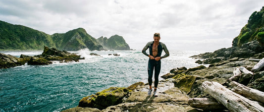 A swimrun athlete in a wetsuit climbing out of the ocean onto rocky coastal terrain with dramatic New Zealand headlands in the background.